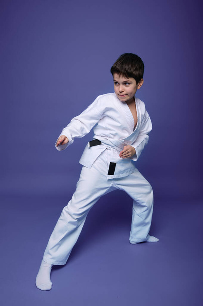 Oriental martial arts. Full length portrait of an aikido fighter, Caucasian 10 years old boy in kimono improves his fighting skills, isolated on violet background with copy space - Photo, Image