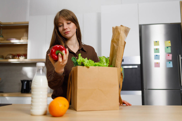 Mulher bonita desembalando legumes e outros alimentos de saco de supermercado de papel na mesa da cozinha - Foto, Imagem