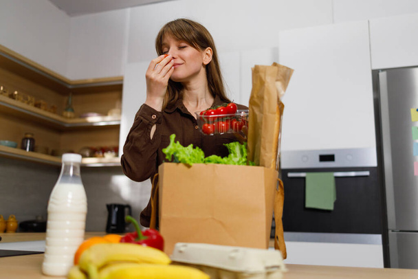 Žena v kuchyni vybalí papírový sáček s potravinami ze supermarketu a očichá čerstvá rajčata - Fotografie, Obrázek