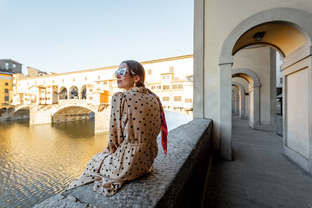 Woman enjoys beautiful view on the old town, sitting on the riverside near famous old bridge in Florence on sunny morning. Stylish woman visiting italian landmarks, traveling Italy - 写真・画像