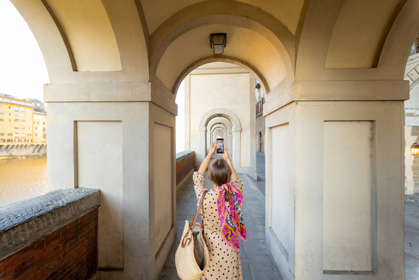 Mujer joven fotografiando por teléfono hermoso arco, visitando la famosa ciudad italiana de Florencia. Turista femenina disfruta de la antigua arquitectura italiana. Mujer vestida al estilo italiano con colorida bufanda en el pelo - Foto, imagen