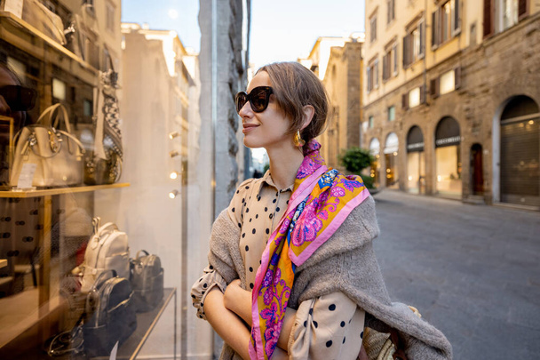 Beautiful woman looks on a showcase with bags and accessories of clothes shop in old town of in Florence city in Italy. Stylish woman wearing colorful shawl in retro italian style - Photo, Image