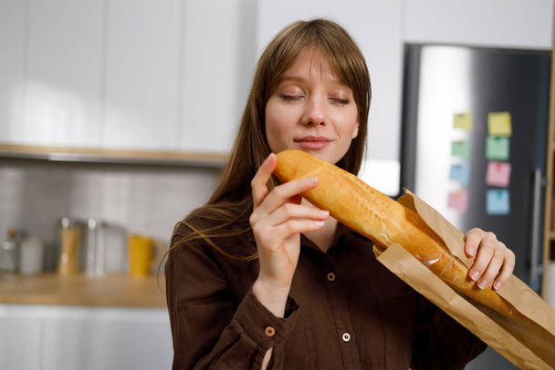 Γυναίκα μυρίζει μια φρεσκοψημένη γαλλική μπαγκέτα ενώ στέκεται στην κουζίνα - Φωτογραφία, εικόνα