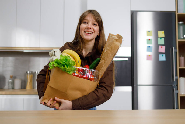 Joven alegre sosteniendo una pesada bolsa de compras de papel llena de comestibles - Foto, imagen
