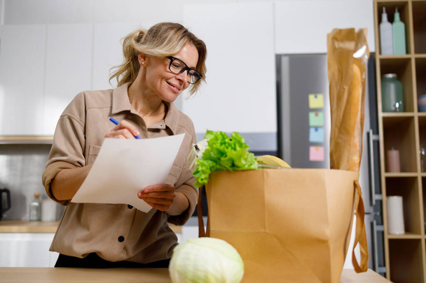 gelukkig volwassen vrouw in keuken holding shopping lijst op zoek door middel van items - Foto, afbeelding