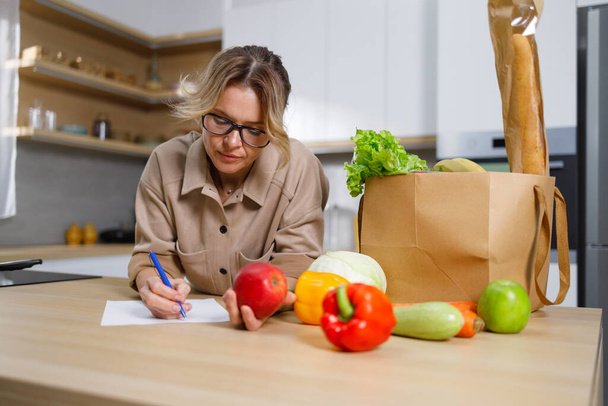 Mature housewife writes down expenses for purchased food. Woman in the kitchen and a paper bag with grocery - Photo, Image