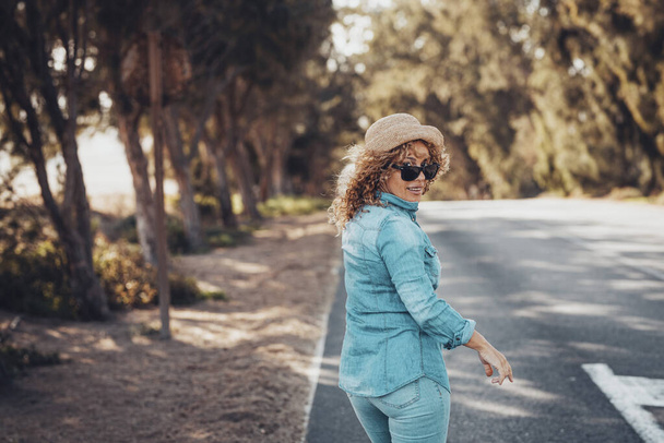 Щаслива жінка дивиться ззаду на камеру і посміхається ходити по дорозі з деревами з обох сторін. Турист з джинсовим повсякденним одягом і солом'яним капелюхом насолоджується активним відпочинком на свіжому повітрі. Копіювати простір
 - Фото, зображення
