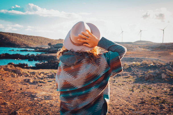 Vista posterior del estilo de vida de la mujer viajera sosteniendo sombrero y admirando un paisaje escénico en el fondo con turbinas de viento y playa del océano. Concepto de personas libres y vida de viaje. Ocio al aire libre - Foto, Imagen