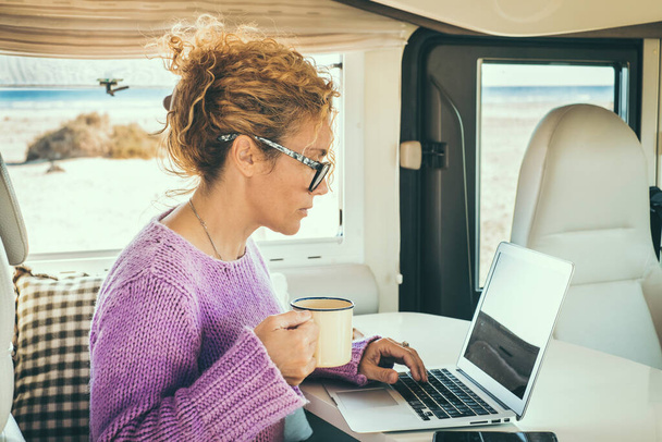 Yetişkin bir kadın karavanın içinde dizüstü bilgisayar kullanıyor. Yolculuk sırasında dijital göçebe yaşam tarzı ve kablosuz internet bağlantısı kavramı. Minibüs hayatı ve akıllı iş kavramı. Sevimli kadın bilgisayarda yazıyor. - Fotoğraf, Görsel
