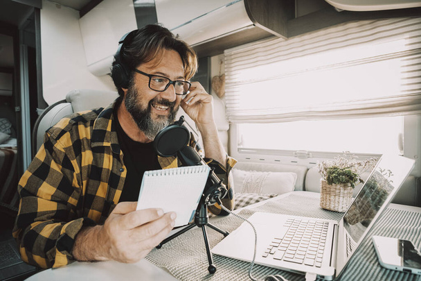 Moderne volwassen man werkt met laptop en microfoon in een camper in digitale nomade baan levensstijl. Technologie en reizen. Alternatieve vrijheid kantoor concept met nieuwe activiteit en vrije mensen - Foto, afbeelding