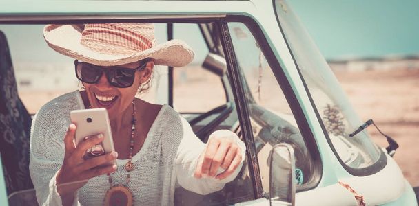 Mutlu genç bir kadın klasik bir minibüste otururken cep telefonuyla iyi eğlenceler. Seyahat ve özgürlük insanların yaşam tarzı. Yaz tatili kavramı. Güzel bir kadın hayattan ve hücresel bağlantıdan hoşlanır. - Fotoğraf, Görsel