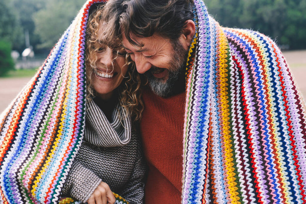 Overjoyed adult caucasian couple under a colorful cover laugh and have fun together with love and friendship. Young mature people in outdoor leisure romantic activity. Man and woman laughing a lot - Foto, Imagen