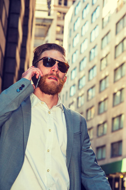 American businessman with beard, mustache traveling, working in New York, wearing cadet blue suit, white undershirt, sunglasses, walking through crowded high building street, talking on cell phone - Фото, изображение