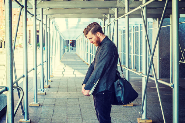 Lost. American student with beard, mustache studying in New York, shoulder carrying leather bag, holding laptop computer, standing on sidewalk bridge, lowering head, sad.  - 写真・画像