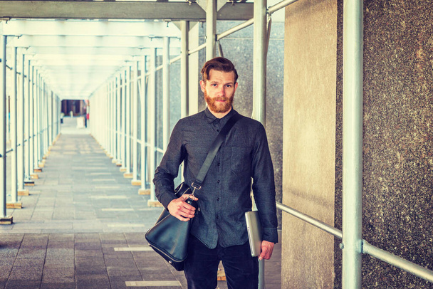 Étudiant américain avec barbe, moustache voyageant à New York, portant une chemise noire, un sac en cuir, tenant un ordinateur portable, marchant sur le pont du trottoir. - Photo, image