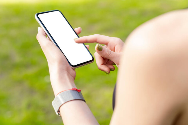 Young caucasian woman wearing sports bra standing on city park, outdoors hands holding phone with mockup white blank display, empty screen for social media. Mobile app tech concept. - Photo, image