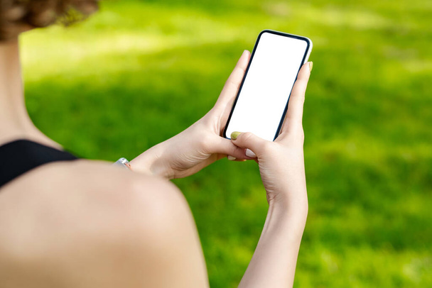 Młoda ruda kobieta w sportowych ubraniach w parku miejskim, na zewnątrz ręce trzymając telefon z makietą biały pusty wyświetlacz, pusty ekran dla mediów społecznościowych. Koncepcja technologii aplikacji mobilnych. - Zdjęcie, obraz