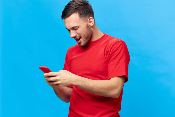 Keskittynyt onnellinen hymyilevä ruskettunut komea mies punaisessa t-paidassa juttelemassa ystävien kanssa käyttäen puhelinta, joka poseeraa sinisellä studiotaustalla. Kopioi tilaa Banner Mockup. Online Ihmiset elämäntapa käsite - Valokuva, kuva