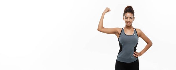 Concepto saludable y fitness: retrato de una joven y hermosa afroamericana que muestra su fuerte músculo con una expresión facial alegre y segura. Aislado sobre fondo blanco estudio
 - Foto, imagen