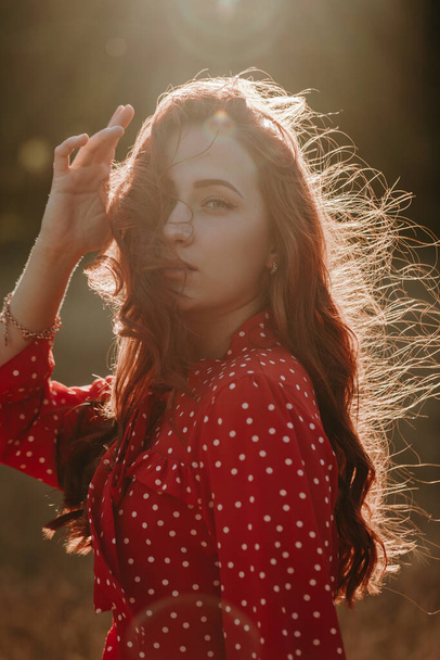Portait de belleza joven pelirroja con largo pelo rizado grueso vestida con camisa roja vestido con puntos blancos posando en una cámara a la luz del sol - Foto, imagen