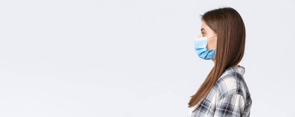 Coronavirus outbreak, leisure on quarantine, social distancing and emotions concept. Profile of serious-looking young pretty woman in medical mask standing in line, white background. - Photo, image