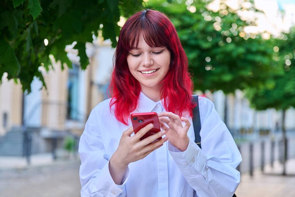 Beautiful smiling teenage girl looking at phone on city street. Female teenager with red dyed hair with backpack using smartphone. Lifestyle, technology, communication, youth, young people - Zdjęcie, obraz