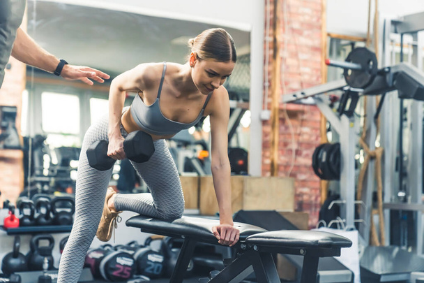 Strong fit sporty caucasian woman in her 20s lifting weights with one hand and leaning on a bench with the other. Gym facility concept. High quality photo - Photo, Image