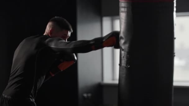 Boxer atletico che indossa vestiti sportivi che colpiscono il sacco da boxe in palestra. Mid shot - Filmati, video