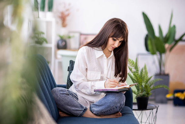 An attractive dark-haired adult girl in a stylish white shirt sits relaxed on a soft blue sofa in her cozy bright apartment, and enthusiastically writes something in her diary - Фото, изображение