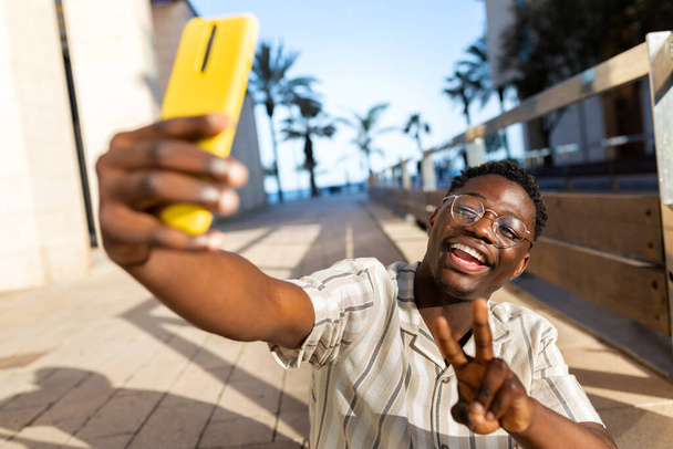 Young black man taking selfie using phone making peace sign. Happy, African american male having fun. Social media concept. - Photo, Image