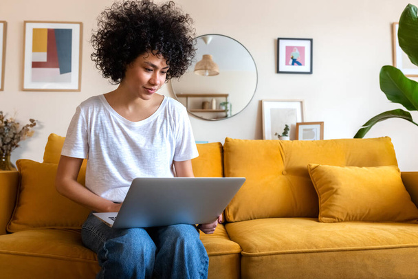 Nuori afroamerikkalainen nainen istuu sohvalla kotona kannettavan tietokoneen ääressä. Kopioi tilaa. Teknologian ja elämäntapojen käsitteet. - Valokuva, kuva