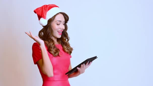 Uma mulher bonita fica em um chapéu de Natal com um tablet, mostra emoções risonhas em um fundo branco. Feliz Ano Novo. - Filmagem, Vídeo