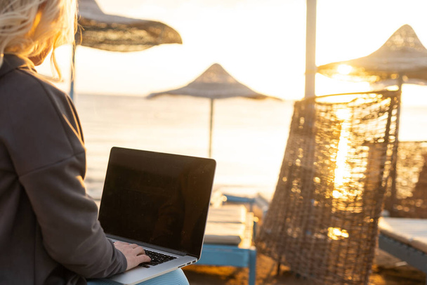 Close-up beeld van een vrouw die op het zandstrand zit en werkt aan een laptop, codeert of e-mails beantwoordt - Foto, afbeelding