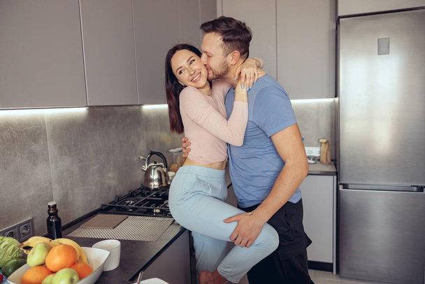 Kaukasischer schöner Mann küsst schöne Frau, während Paar morgens in der Küche steht. - Foto, Bild