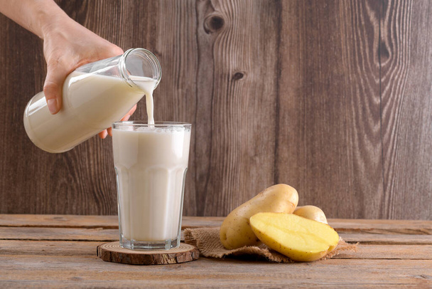 Vegan potato milk is pouring into a glass and potato tubers on rustic wooden background. Plant based alternative milk replacer and lactose free, Trendy healthy vegetarian and vegan drink concept - Photo, Image