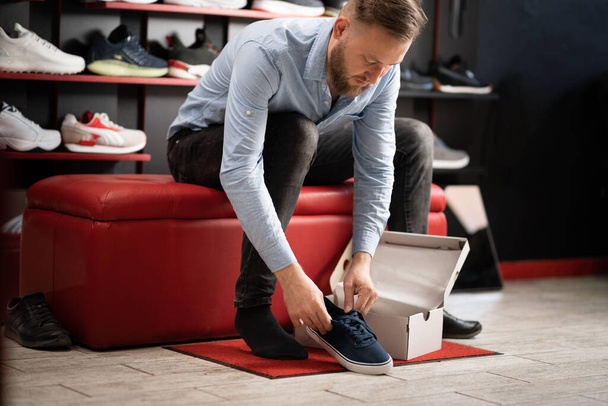caucasian bearded man chooses hiking shoes while sitting in fitting room of modern sports store. Puts on shoes and selects the appropriate style and size. copy space. - Фото, изображение