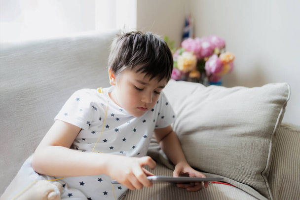 High key portrait kid playing game on tablet sitting on sofa with light shining from window, Young boy  playing games on internet, Cinematic portrait Child doing homework online at home - Photo, Image