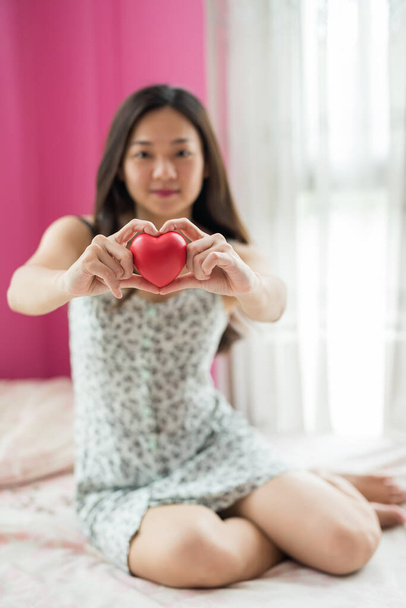 Asiático bonito jovem mulher segurar coração vermelho com 2 mãos gesto na cama. Ela está apaixonada no dia dos namorados.. - Foto, Imagem