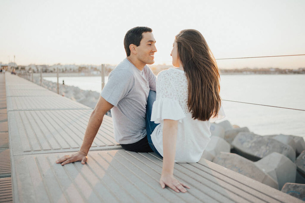 Un uomo ispanico e una ragazza bruna sorridente sono seduti e si fissano a vicenda su un frangiflutti in Spagna la sera. Un paio di turisti in un appuntamento al tramonto a Valencia. - Foto, immagini