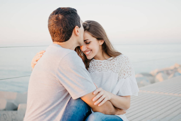 Ein hispanischer Mann küsst abends auf einem Wellenbrecher in Spanien seine lächelnde brünette Freundin auf die Stirn. Ein paar Touristen bei einem Date bei Sonnenuntergang in Valencia. - Foto, Bild