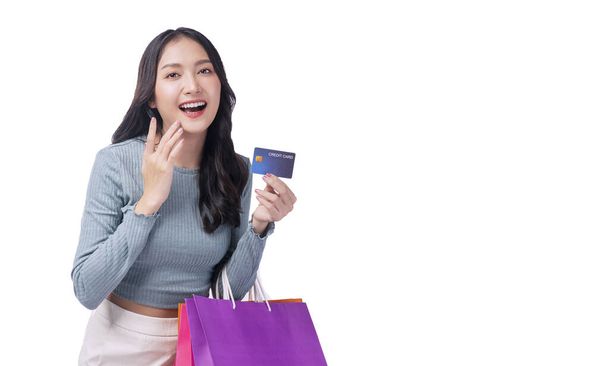 Возбужденный азиат показывает кредитку, держащую сумки с покупками на белом фоне. Веселая девушка-покупатель с помощью пластиковой карты оплаты проведения покупки бумажные пакеты на продажу на изолированном фоне - Фото, изображение