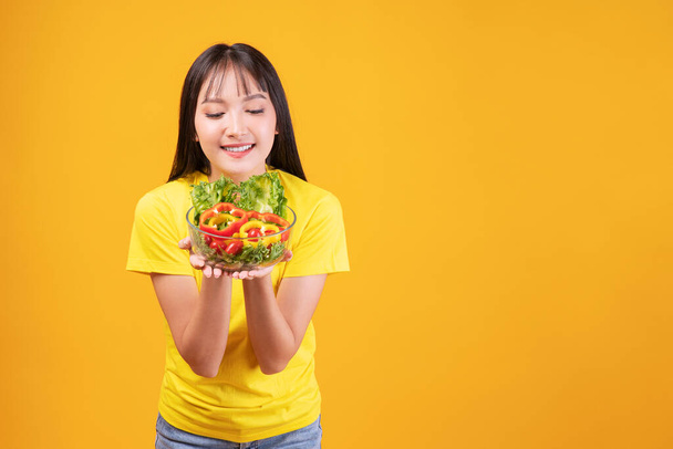Cheerful Asian woman slim body holding glass bowl with organic fresh vegetables for good health diet standing over isolated yellow background. Fit body young girl Health care lose weight food concept. - Foto, afbeelding