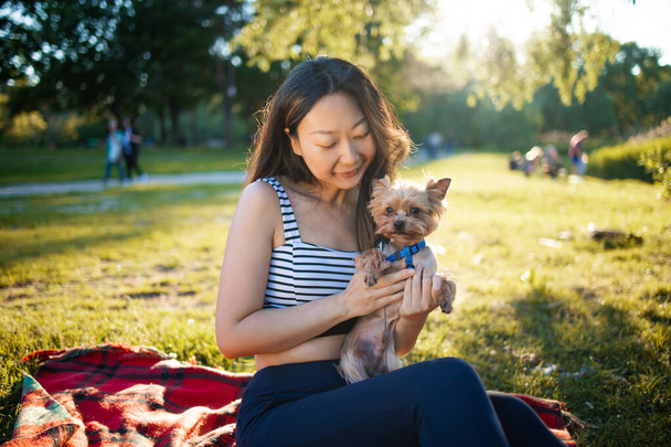 Νεαρή όμορφη χαμογελαστή Ασιάτισσα που κρατάει το σκύλο της στη φύση στο καλοκαιρινό πάρκο το ηλιοβασίλεμα. - Φωτογραφία, εικόνα