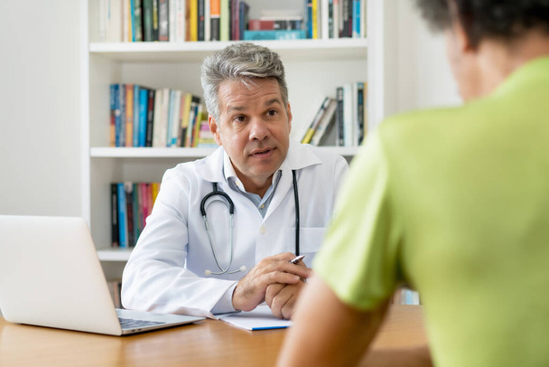 Serious doctor with gray hair talking with patient at office of hospital - Photo, Image