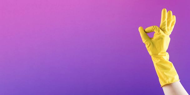 Hand in yellow rubber glove shows OK gesture on purple background closeup. Professional cleaning service. Easy housework and routine chores - Valokuva, kuva