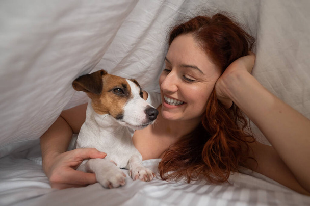 Caucasian red-haired woman sleeps in an embrace with a jack russell terrier dog on a white sheet - Foto, imagen