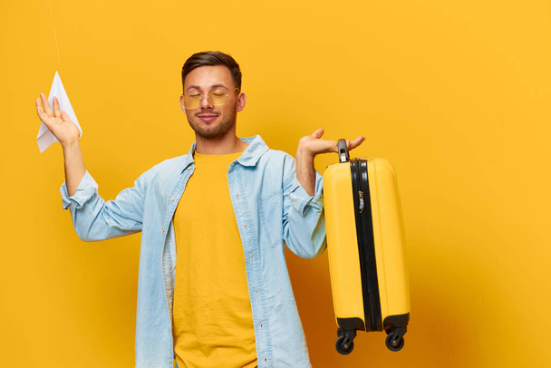 青いシャツのトレンディなアイウェアで幸せな若い日焼けしたハンサムな男をリラックスして,オレンジ色のイエロースタジオの背景に隔離された紙飛行機のスーツケースを保持します. スペースバナーモックアップをコピーします. 旅のコンセプト - 写真・画像