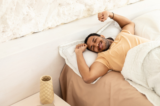 Hombre árabe alegre despertar estiramiento brazos acostado en la cama disfrutando de la mañana después de un buen sueño en el dormitorio moderno en el interior. Hombre despertando después del descanso en casa. Concepto de sueño saludable - Foto, Imagen
