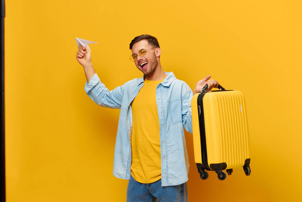 Nauttinut iloinen ruskettunut komea mies sininen paita trendikäs silmälasit heittää paperin lentokone pidä matkalaukku poseeraa oranssi keltainen studio tausta. Kopioi tilaa Banner Mockup. Matkan käsite - Valokuva, kuva