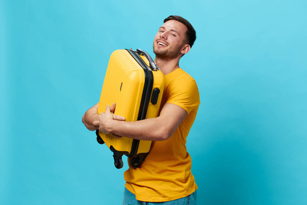 Sarı tişörtlü, mutlu, bronzlaşmış yakışıklı adam mavi stüdyo arka planında izole edilmiş bir şekilde duran bavulu tutmak için kameraya bak. Uzayın afiş modelini kopyala. Seyahat konsepti - Fotoğraf, Görsel